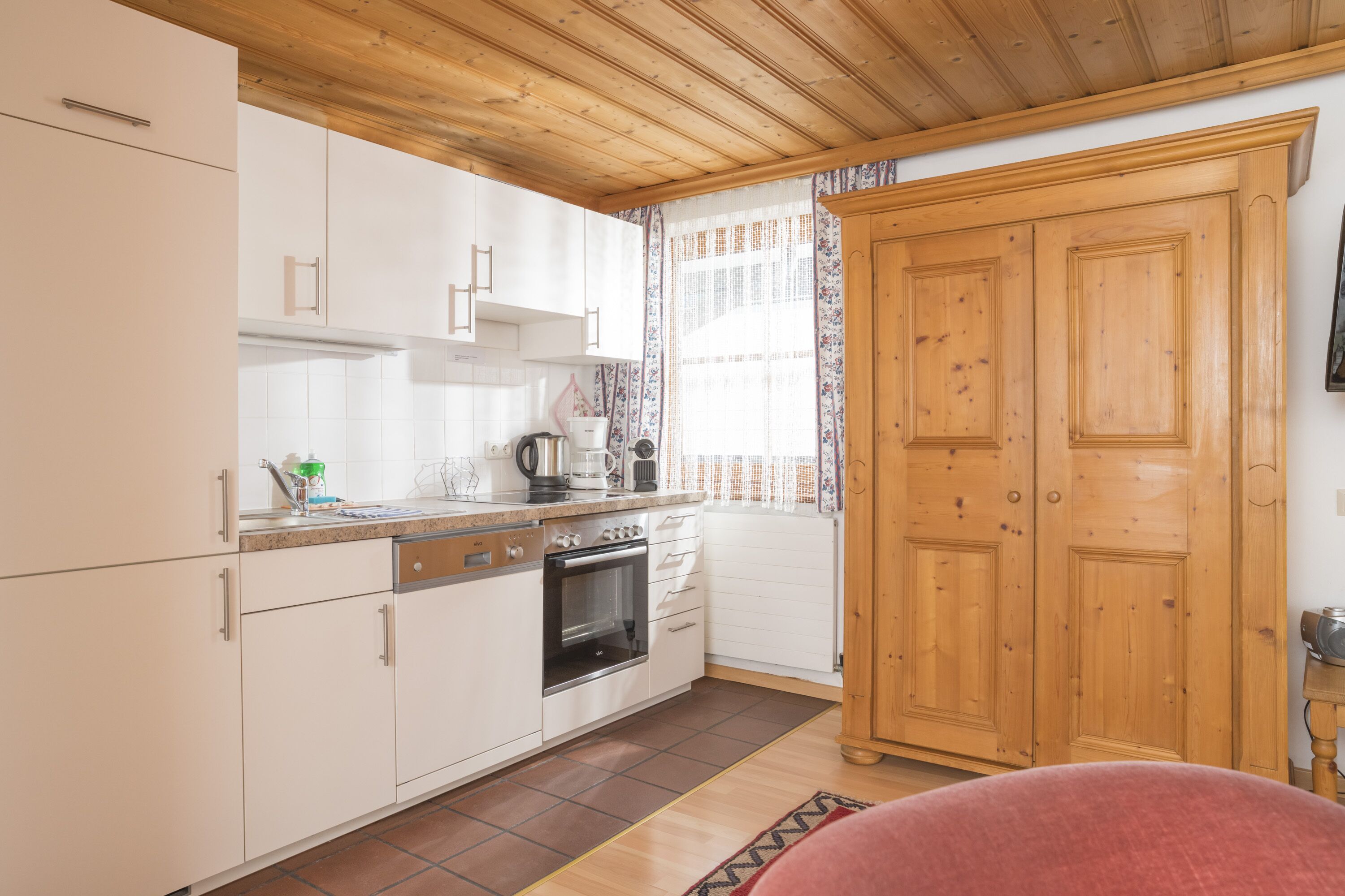 A kitchen with white cabinets, a wooden ceiling, a large wooden wardrobe, and a window with curtains.