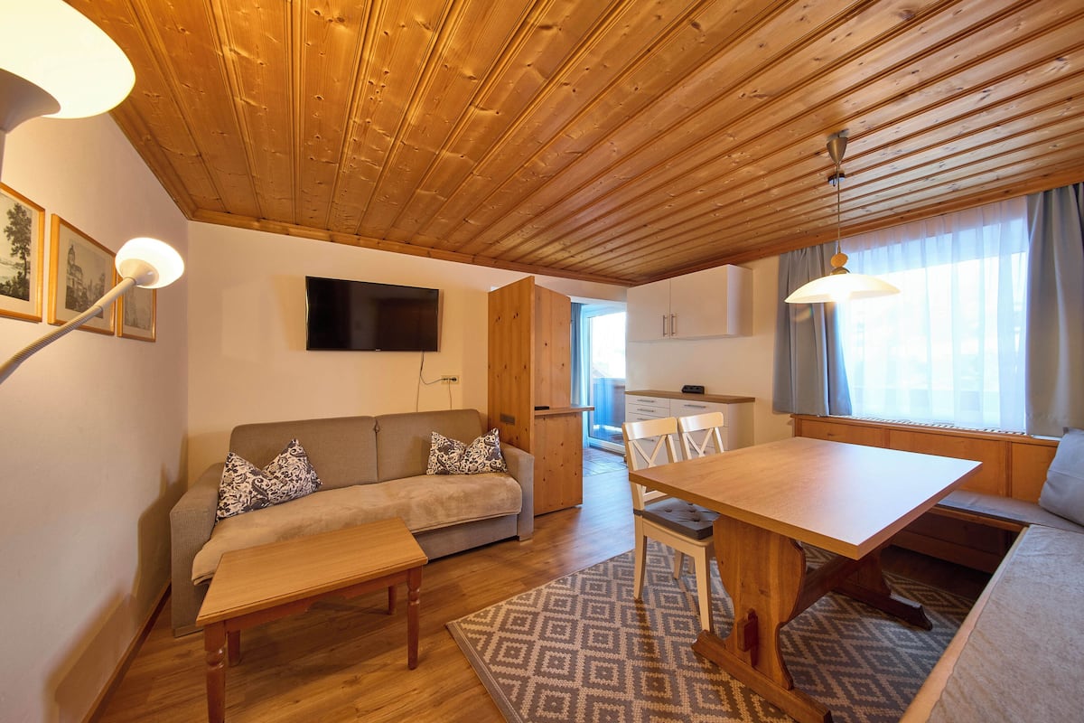 Ein gemütliches Zimmer mit Holzboden, einer Couch, einem Esstisch und einem an der Wand befestigten Fernseher.