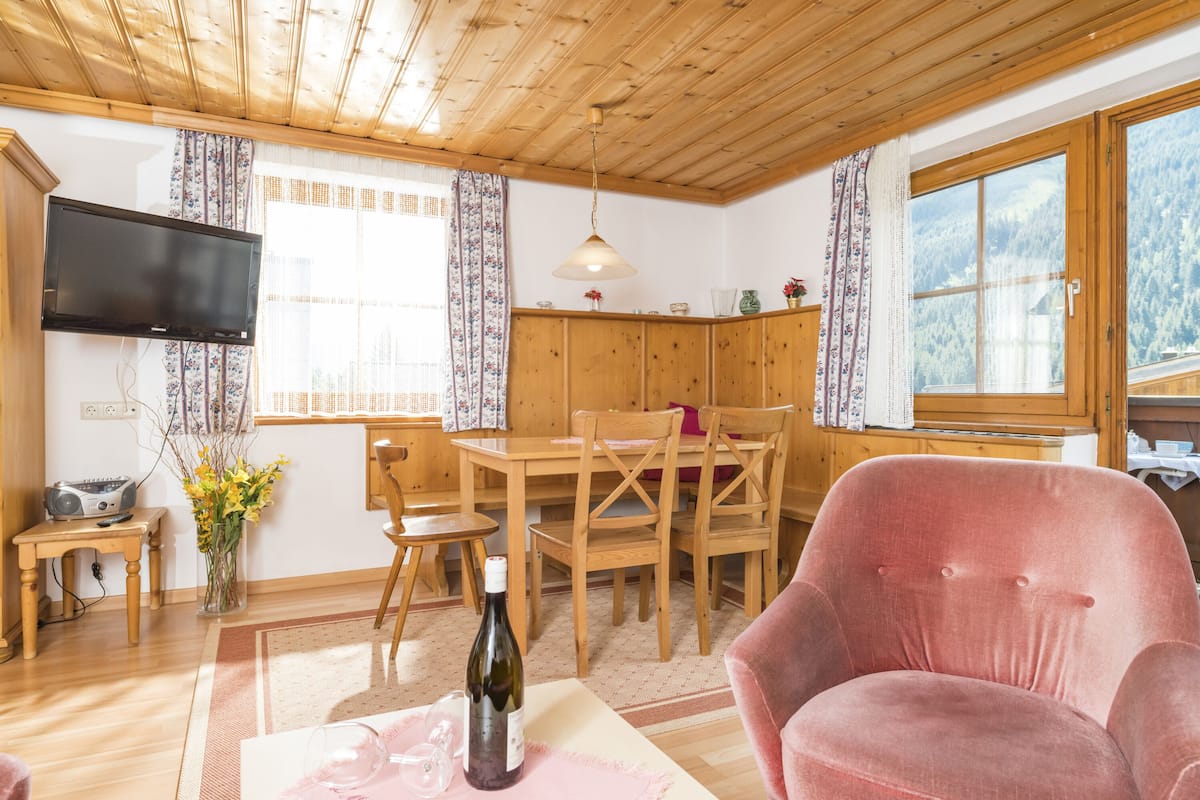 Ein gemütliches Zimmer mit Holzmöbeln, einem Esstisch, Stühlen, einem Fernseher und einem Fenster mit Blick auf einen Wald.