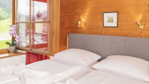 A wooden cabin room with two beds, a window with a view of a green landscape, and a framed picture on the wall. - Appartements Bernhofer  (Saalbach-Hinterglemm)