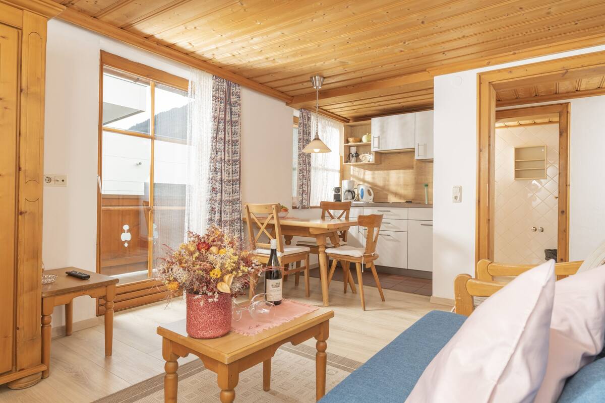 Ein gemütliches Zimmer mit Holzbalkendecke, einem Essbereich mit Stühlen und Tisch, einer Küchenzeile mit Schränken und einem großen Fenster mit Vorhängen.