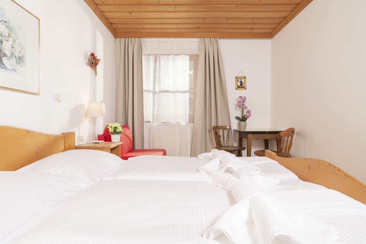 Ein Hotelzimmer mit Holzbalkendecke, einem großen Bett mit weißer Bettwäsche, einem kleinen Schreibtisch mit Lampe, einem Sessel und einem Fenster mit Vorhängen.