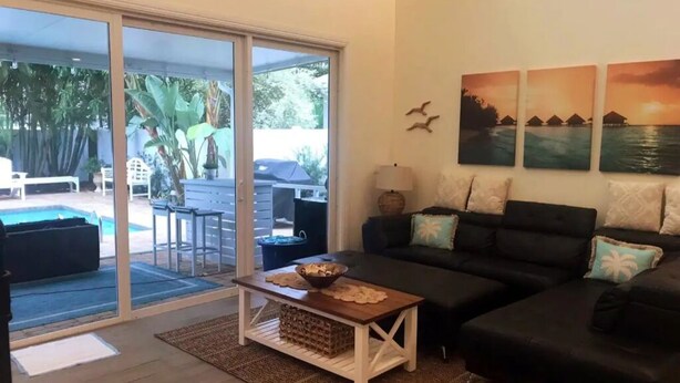 Ein Wohnzimmer mit schwarzem Sofa, einem Holztisch und einer Glastür, die zu einem Außenbereich mit Pool und Terrassenmöbeln führt.