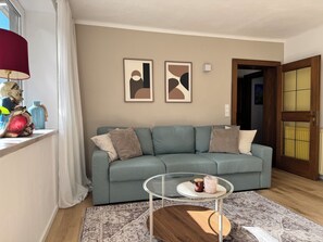 A living room with a sofa, a glass coffee table, and a patterned rug. There are abstract paintings on the wall and a window with curtains. - Edeltraud Vacation Apartment on Lake Millstatt: Hiking Vacations and Much More, Apartment 1 (Radenthein)