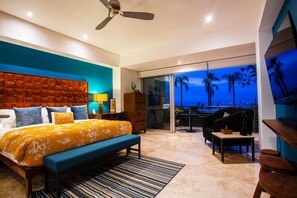 A modern bedroom with a large bed, a desk, a chair, and a balcony with a view of palm trees.