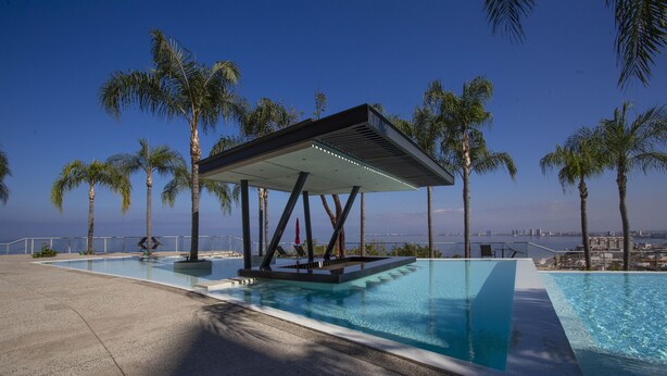 A modern infinity pool with a covered seating area, surrounded by palm trees and overlooking a cityscape. - Fully remodeled 12 stories up! First floor retreat!! (Puerto Vallarta)