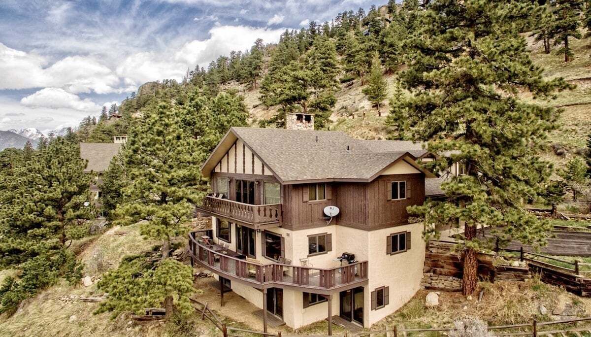 A multi-story house with a balcony, surrounded by trees and a mountainous backdrop.