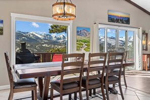 Ein Essbereich mit Tisch und Stühlen, großen Fenstern mit Bergblick, einer Kandelaber und einem Bild an der Wand.