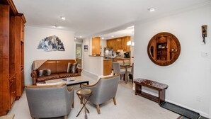 Una sala de estar moderna con un sofá de cuero marrón, sillones grises y una mesita de centro. A la izquierda hay un mueble de madera y, al fondo, una zona de cocina.
