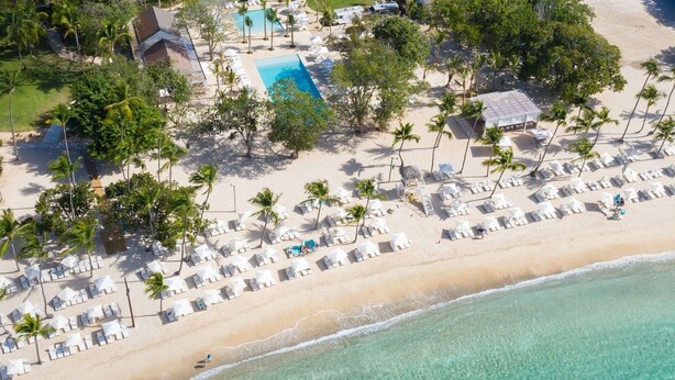 Vue aérienne d’un complexe hôtelier de bord de mer, avec une piscine, des chaises longues et des palmiers.