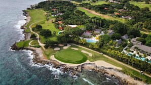 Une vue aérienne d’un parcours de golf situé sur la côte, offrant une vue dégagée sur l’océan, un terrain verdoyant bien entretenu et divers bâtiments et constructions.
