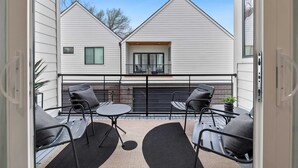 A balcony with two tables and chairs, a small window, and a view of a white building.