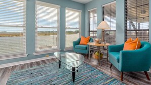 A spacious living room with large windows, teal armchairs, a glass coffee table, and a small round table with a lamp.
