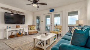 A living room with a TV, ceiling fan, white furniture, and a view through the glass doors.