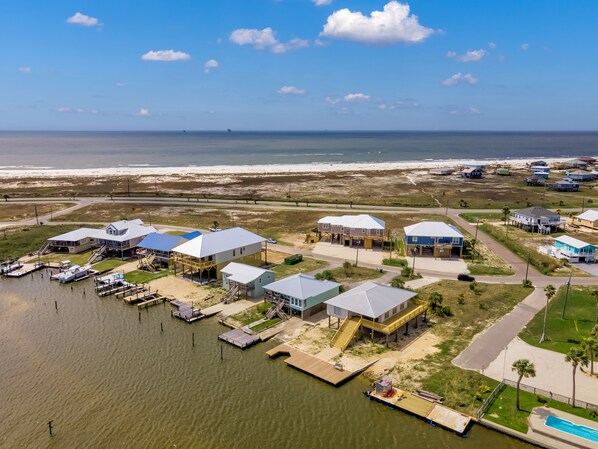 An aerial view of a coastal residential area with houses connected by walkways to the water, a dock with boats, and a beach in the background.