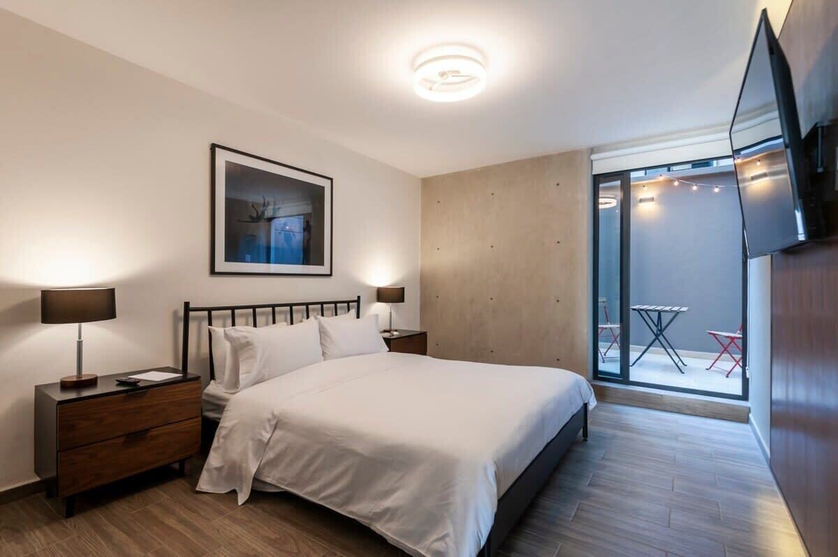 Ein modernes Schlafzimmer mit einem großen Bett, einem Nachttisch mit Lampe, einem gerahmten Bild an der Wand und einer Glasschiebetür zu einem Außenbereich mit Tisch und Stühlen.