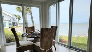 Een eethoek met rieten stoelen en een glazen tafel, met uitzicht op een waterpartij en een gebouw met een balkon.