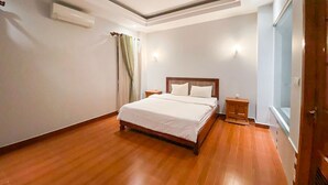 A bedroom with a wooden floor, a bed with white bedding, a nightstand, a window with curtains, and an air conditioning unit on the wall. - Heng Mohasal Apartment (Phnom Penh)