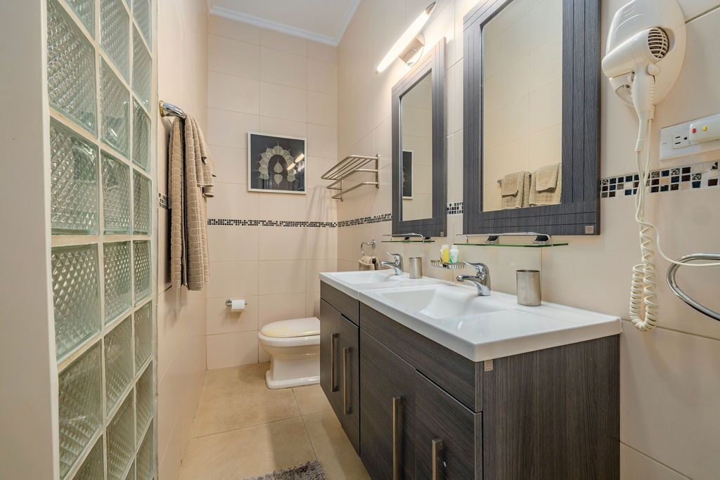 Ein Badezimmer mit einem Doppelwaschbecken, einer Toilette, einer Duschkabine aus Glas und einem an der Wand montierten Fön.