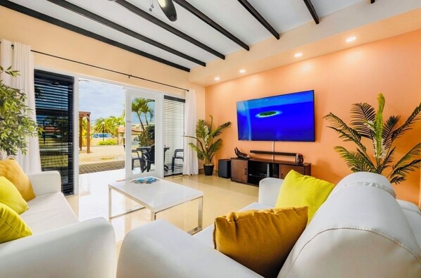 A modern living room with a TV, white sofas with yellow cushions, a coffee table, and potted plants.