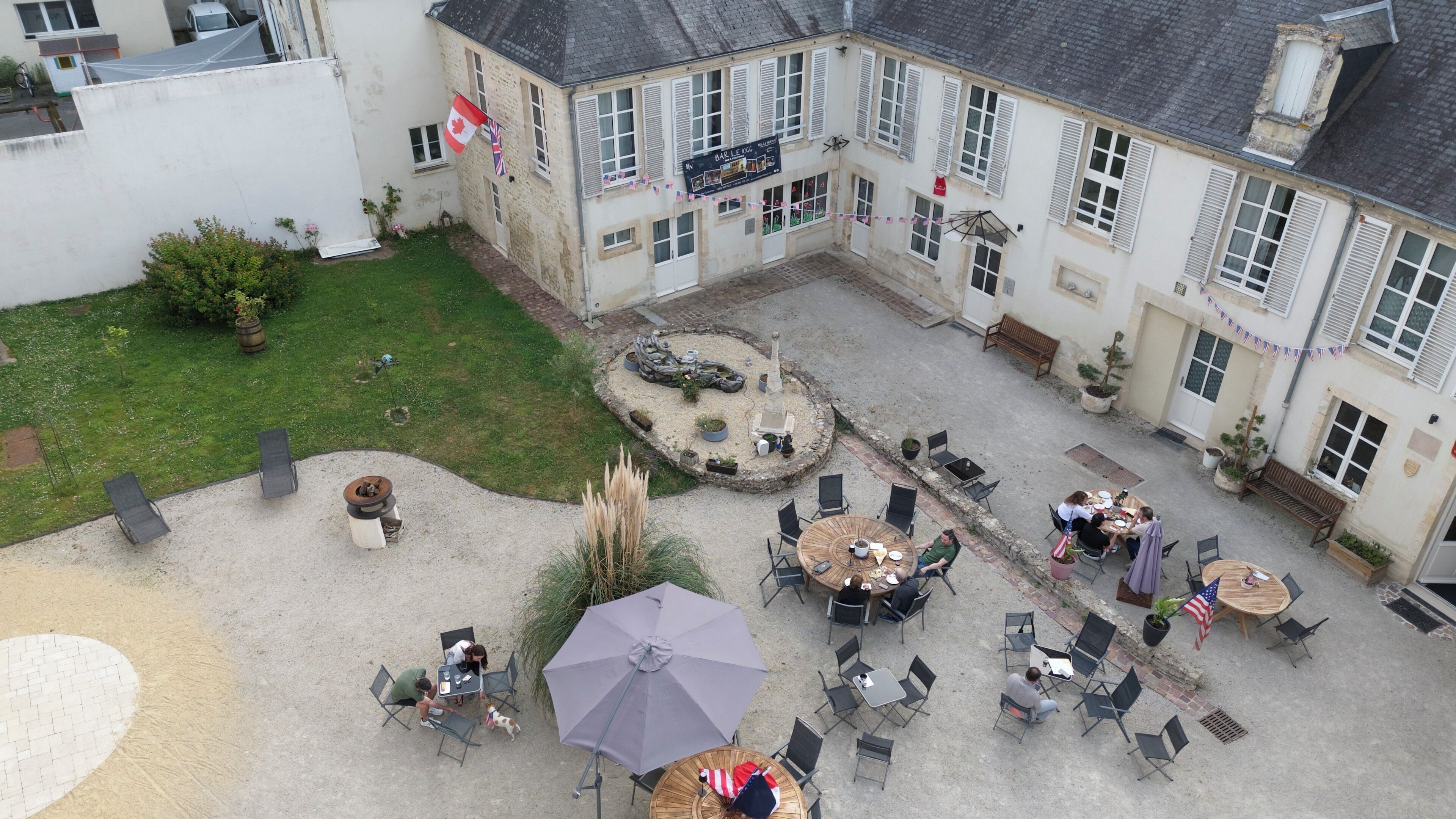 An outdoor courtyard with multiple seating areas, a central garden, and a building with a sign reading "BAR LE DOG".