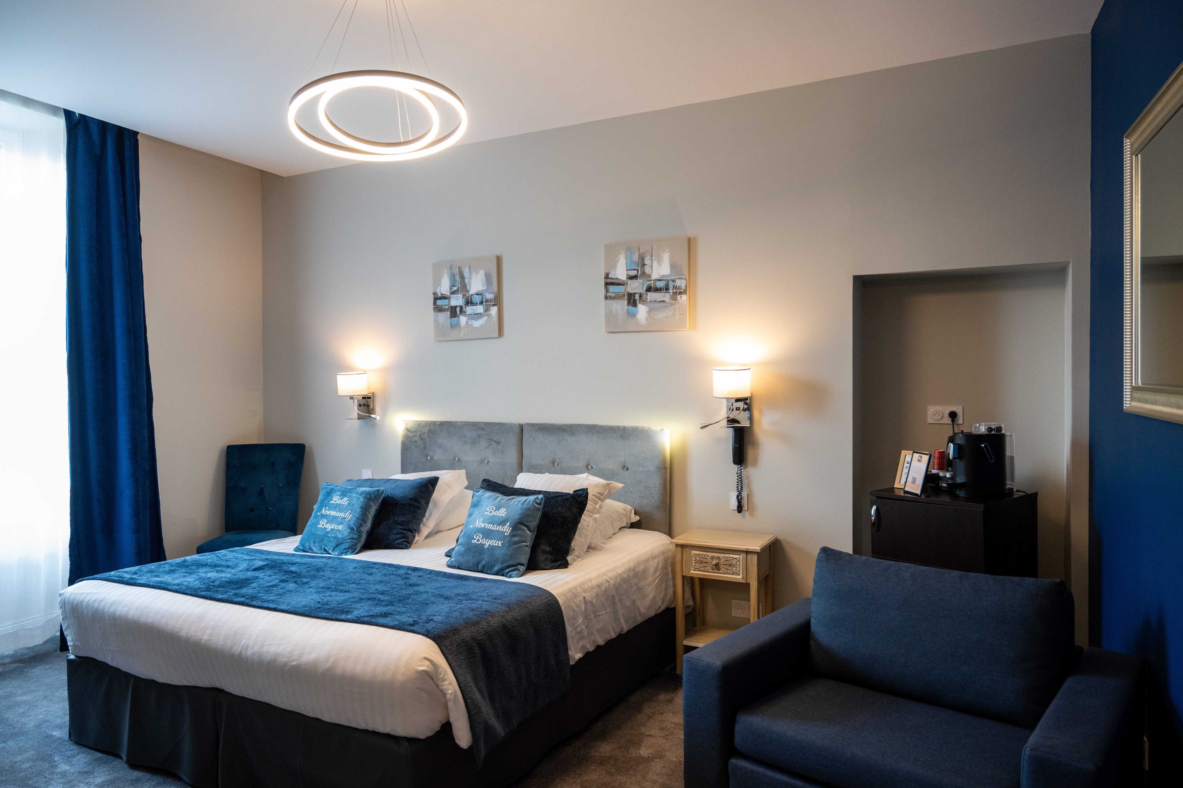 A modern hotel room with a large bed, bedside lamps, a headboard, artwork on the wall, a small table with a coffee machine, and a chair.