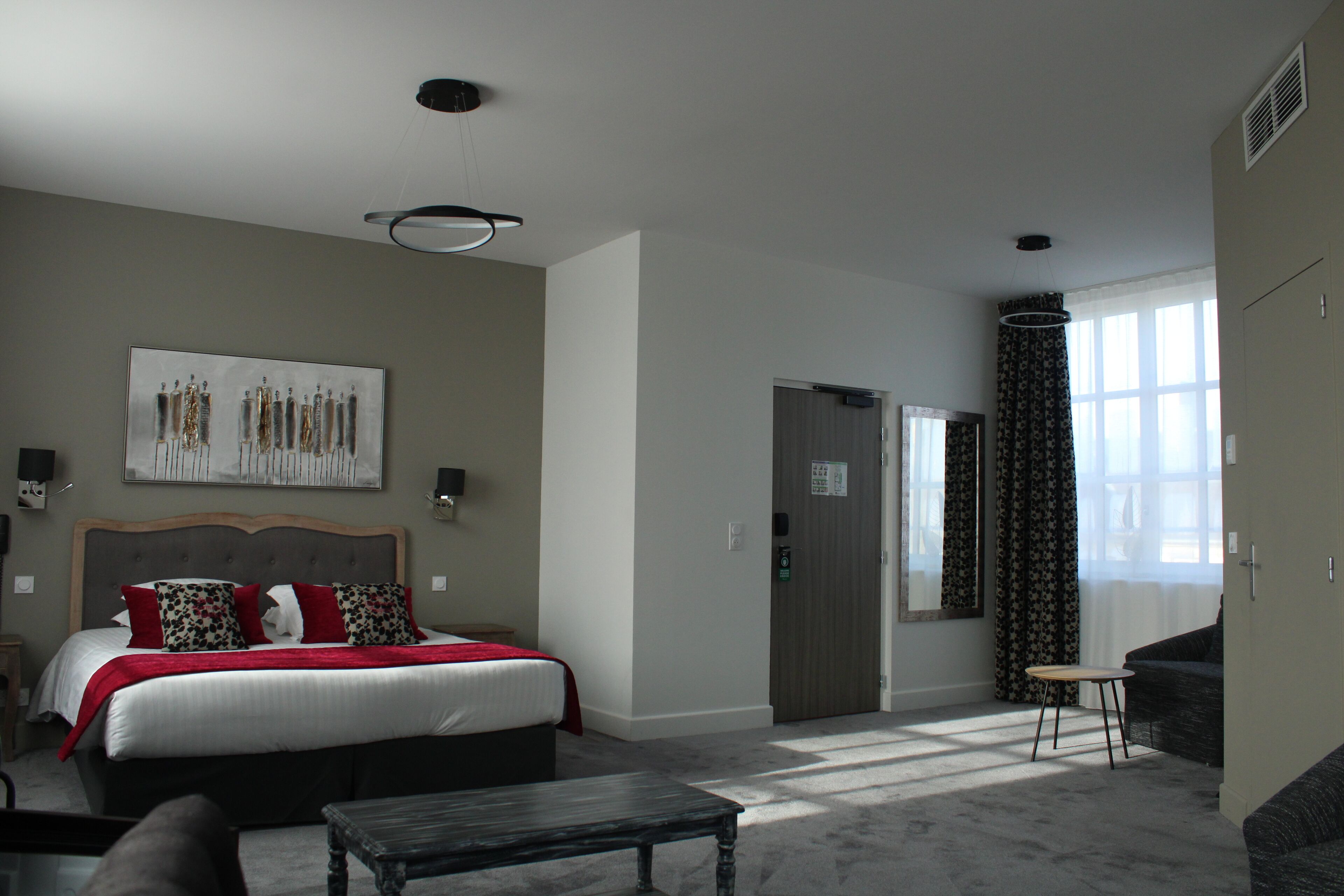 A hotel room with a bed, a small table, a sofa, a painting on the wall, and a window with curtains.