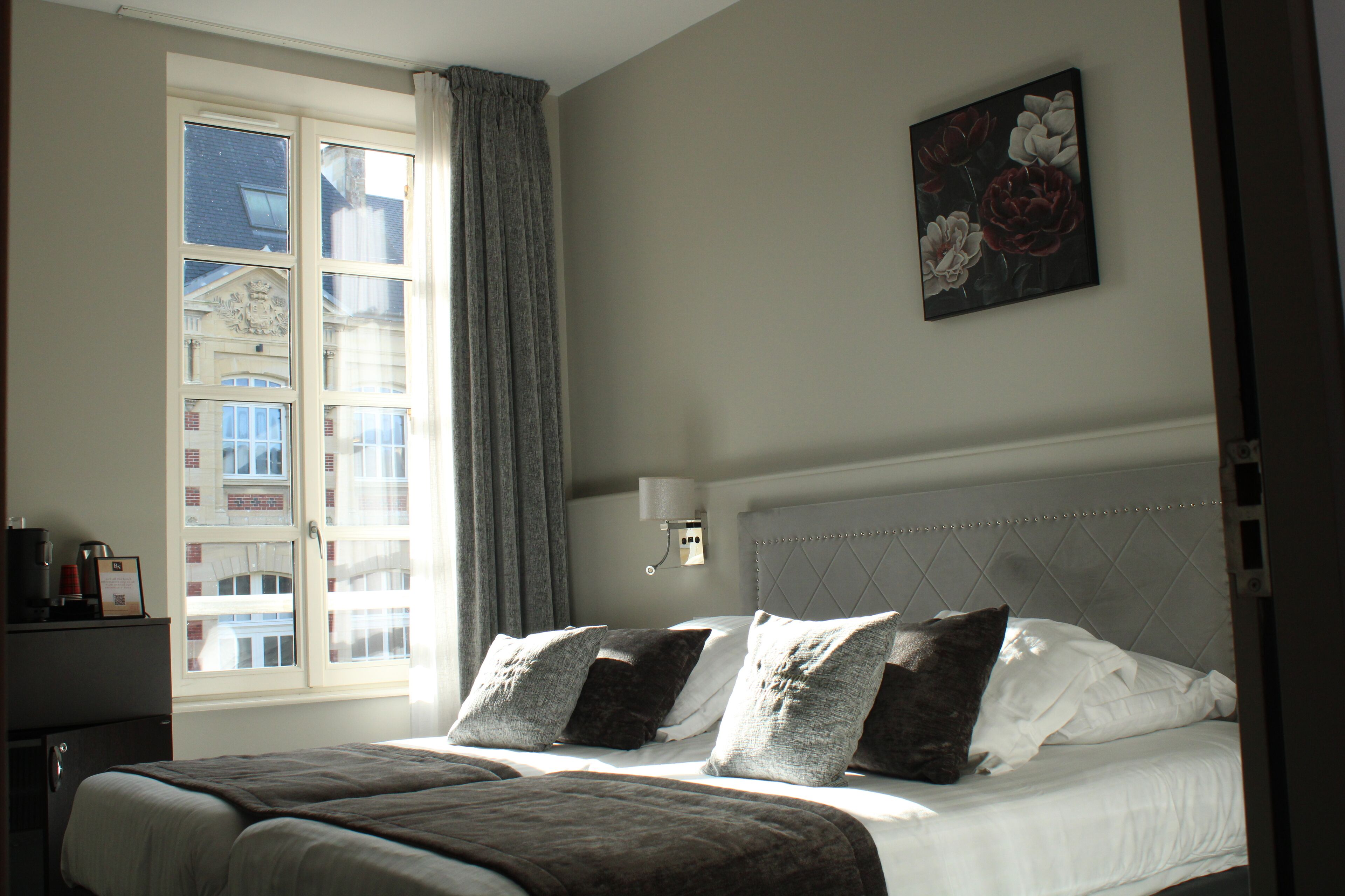 A hotel room with a large bed, a nightstand, a window with curtains, and a wall-mounted lamp.