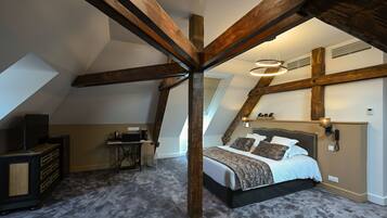 Une chambre avec un plafond en pente, des poutres apparentes, un lit recouvert d’une couvre-lit à motifs, un bureau avec une chaise et une petite table avec une lampe.