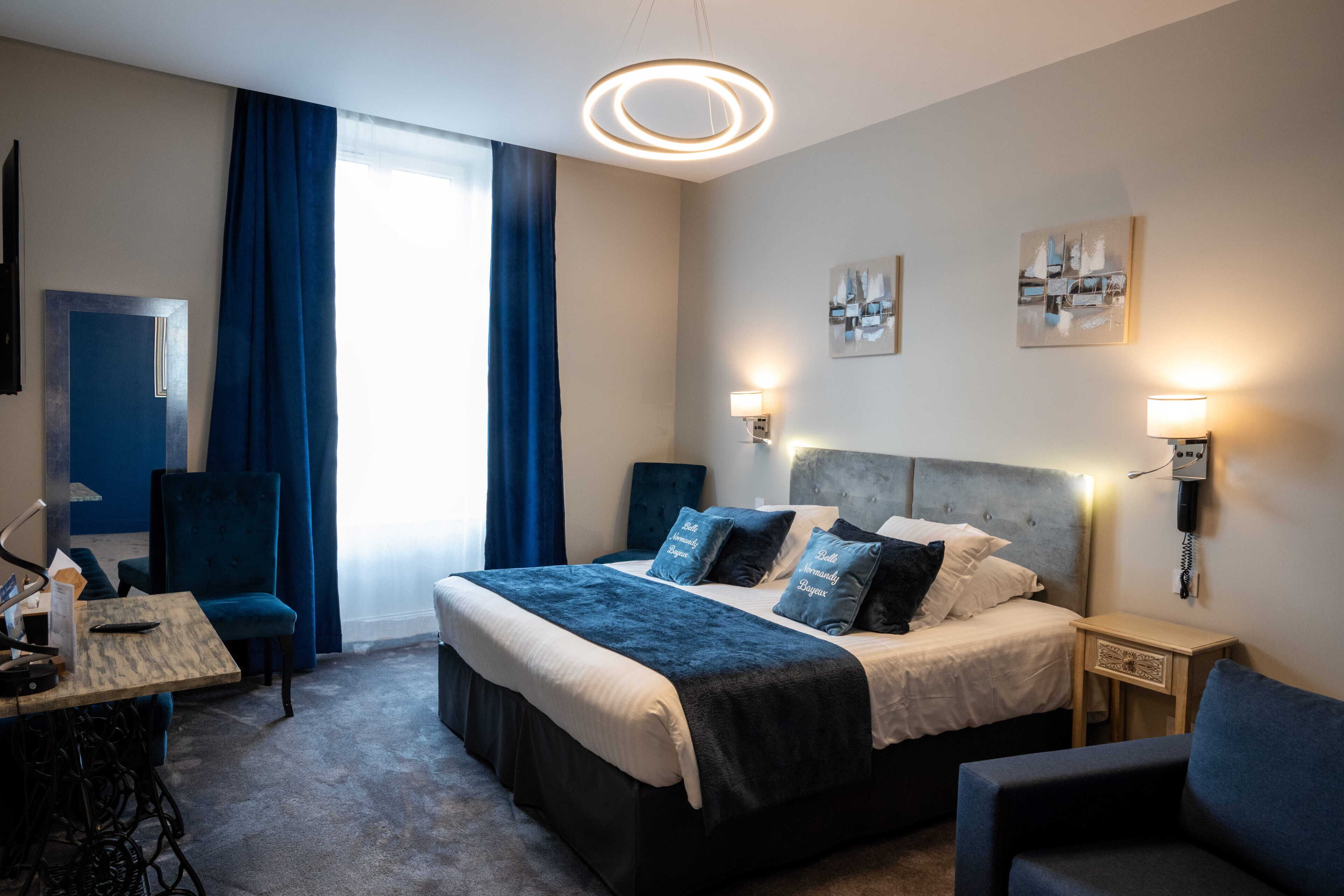 A modern hotel room with a large bed, a desk, a chair, a small table, and a nightstand with a lamp.