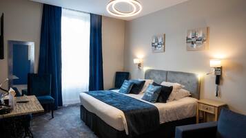 A modern hotel room with a large bed, a desk, a chair, a small table, and a nightstand with a lamp.