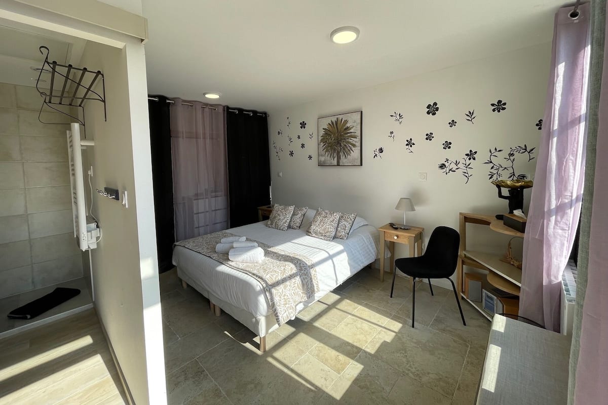 Un dormitorio con cama, una silla, una mesita de noche, una lámpara de pared y un espacio para la ducha.