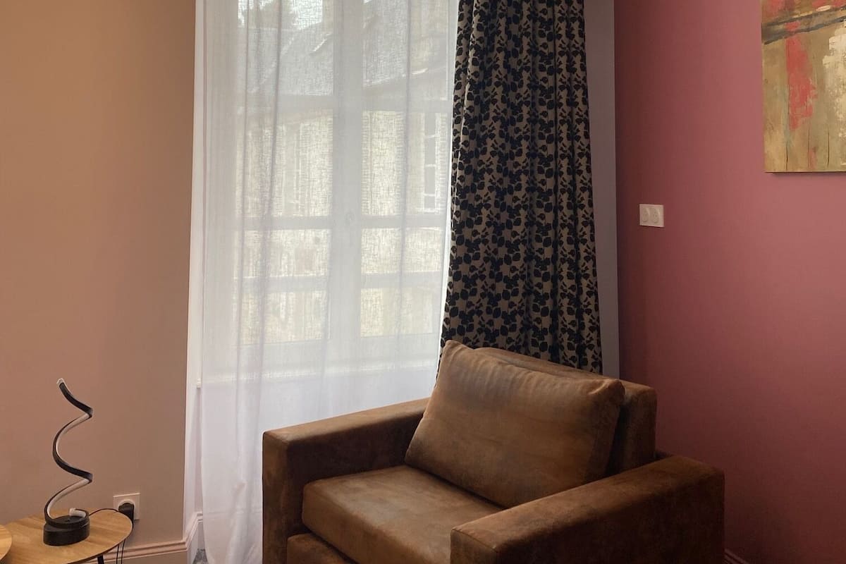 Una habitación con un sofá marrón, una mesita redonda con una lámpara, una ventana con cortinas translúcidas y un cuadro en la pared.