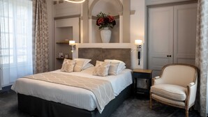 Quarto de hotel com uma cama grande, uma cadeira, mesa de cabeceira, um vaso com flores e um espelho.