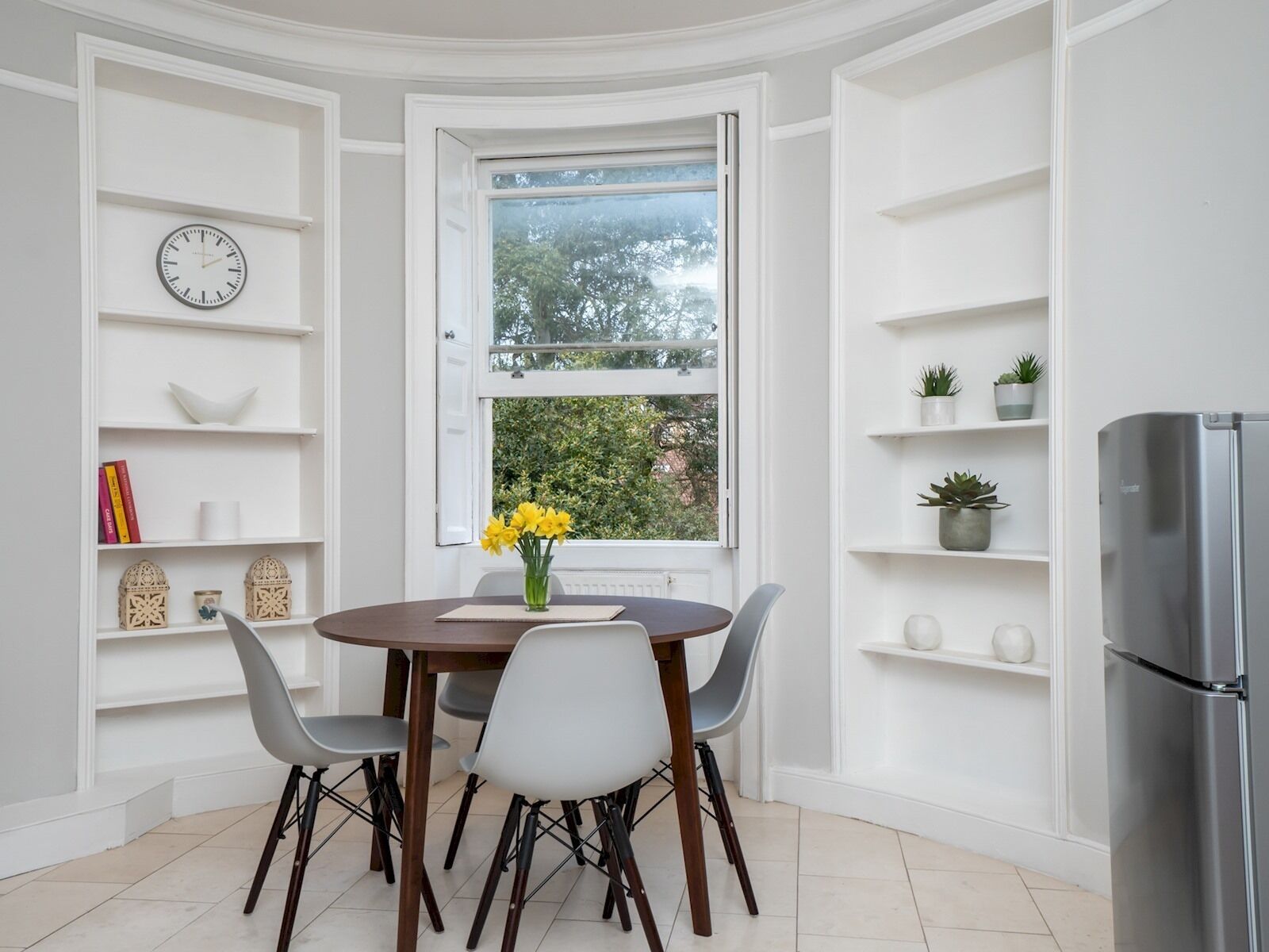 Un coin repas comprenant une table ronde, quatre chaises, une horloge et une fenêtre donnant sur un paysage verdoyant.