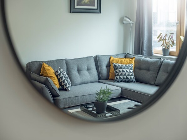 A modern living room with a grey sectional sofa, two yellow and two geometric patterned cushions, a floor lamp, and a potted plant by the window.