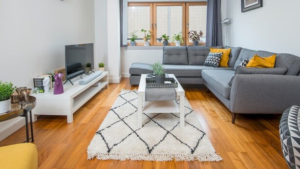 Una sala de estar moderna con un sofá gris, una mesa de centro, un televisor sobre un mueble blanco y una ventana con plantas.