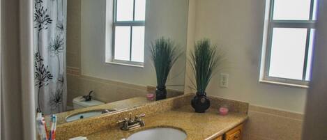 A bathroom with a granite countertop, a sink, a mirror, two potted plants, and a shower curtain with a floral pattern.