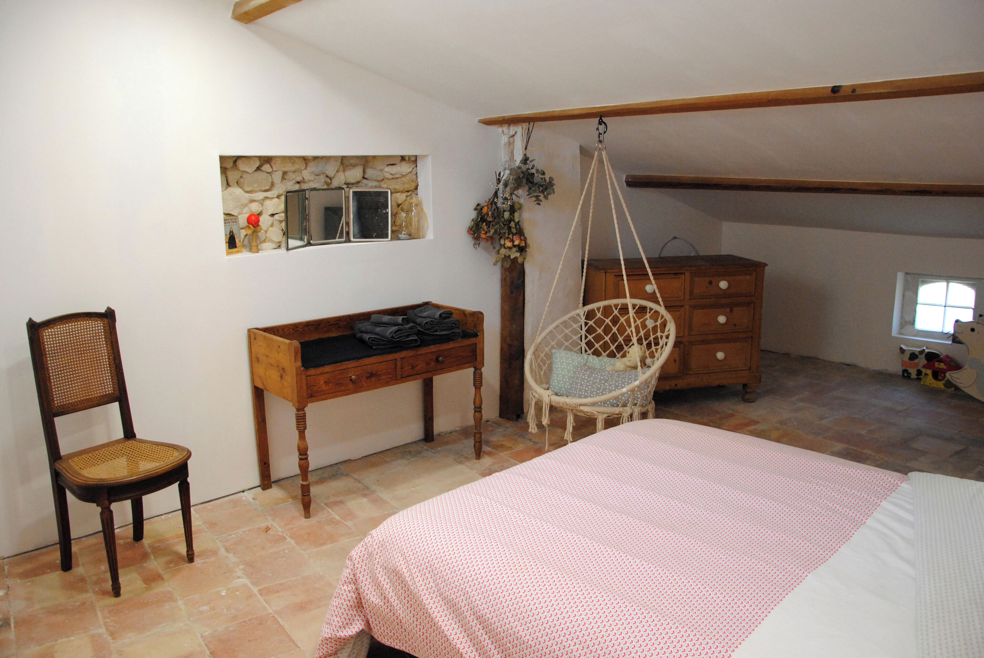Une chambre à coucher avec un lit, une commode en bois, une chaise en osier, un bureau et un mur en pierre.