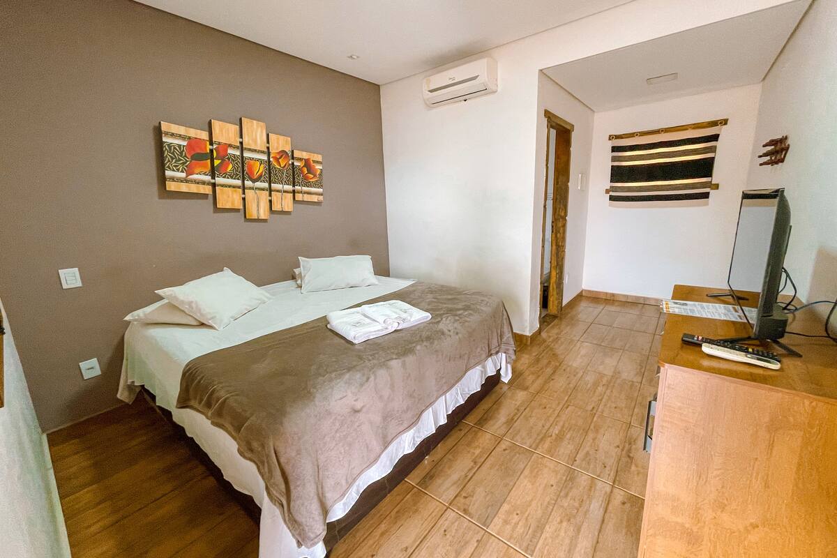 A hotel room with a bed, a desk with a computer and a TV, a painting on the wall, and a door leading to another room.