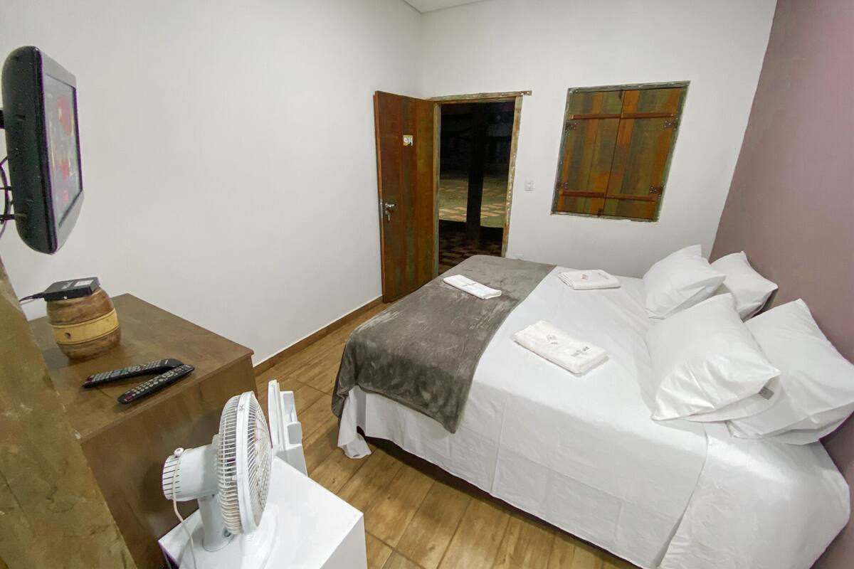 A bedroom with a bed, a bedside table with a fan, a remote control, and a wooden door.