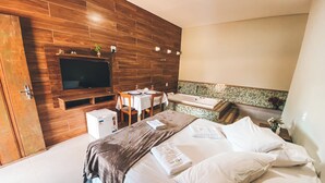 A modern hotel room with a wooden wall, a flat-screen TV, a small refrigerator, a dining area with a table and chairs, and a bathroom with a bathtub. - COMPLEXO JARDIM SECRETO (Santa Barbara)