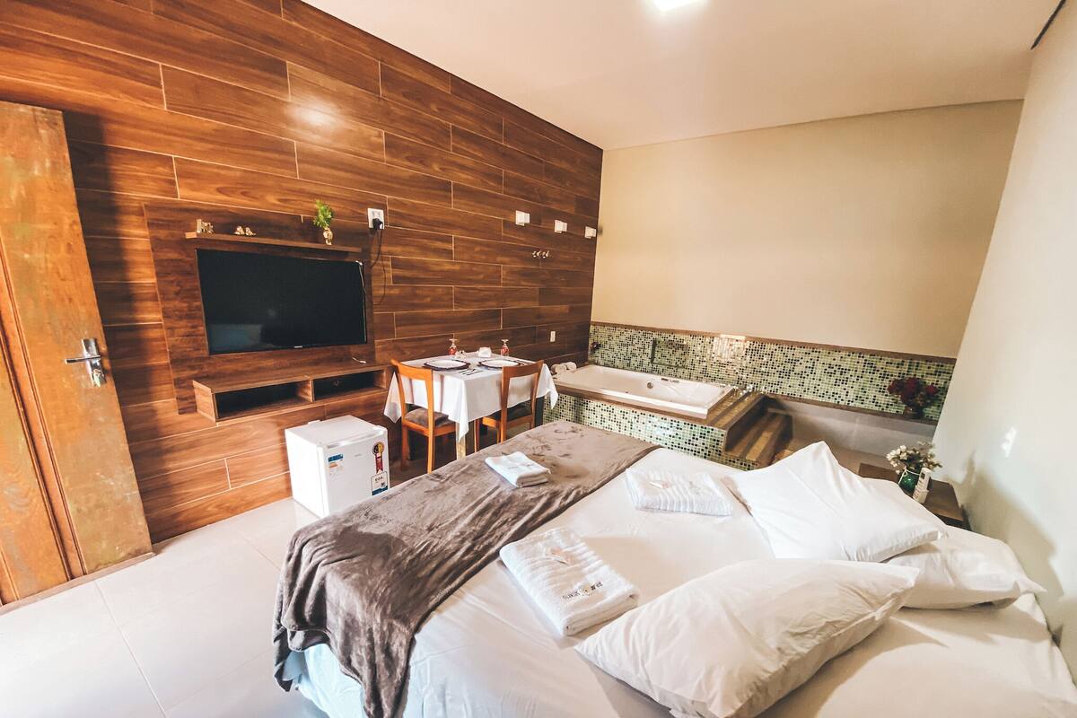A modern hotel room with a wooden wall, a flat-screen TV, a small refrigerator, a dining area with a table and chairs, and a bathroom with a bathtub.