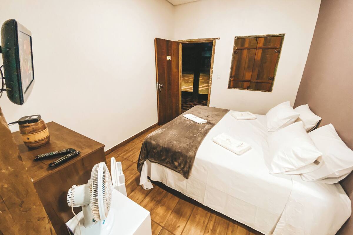 A bedroom with a bed, a bedside table with a fan, a remote control, and a wooden door.