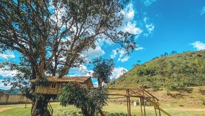 A treehouse with a wooden deck and a slide, surrounded by lush greenery and a clear sky. - COMPLEXO JARDIM SECRETO (Santa Barbara)
