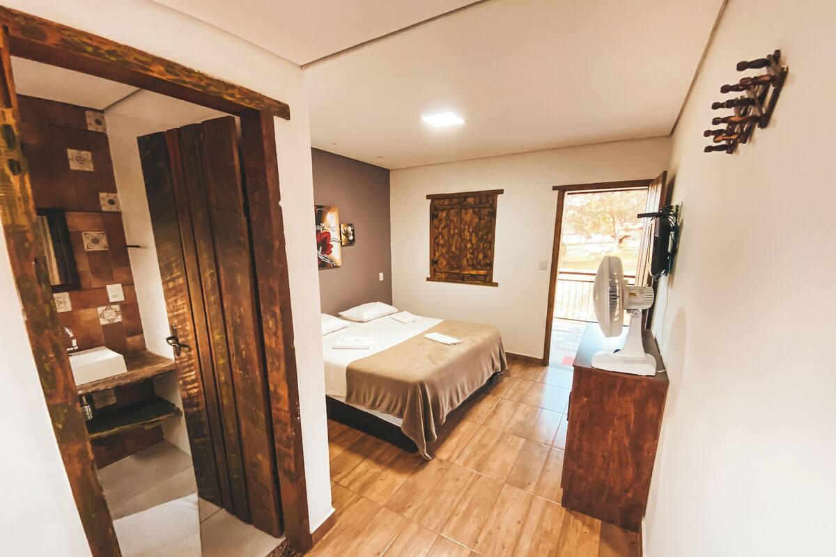 A compact bedroom with a wooden floor, a bed with a beige bedspread, a small bedside table, a wall-mounted fan, and a window with a view.