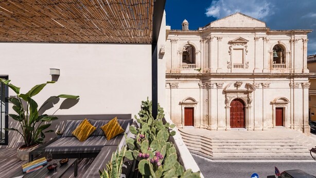 A modern outdoor seating area with a cushioned bench and potted plants, adjacent to a historic building with classical architecture. - Casa San Lio (Noto)