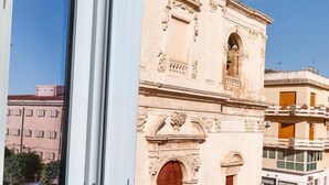 A window with a view of a historic building with ornate architecture, a clear blue sky, and a magazine on the windowsill. - Casa San Lio (Noto)