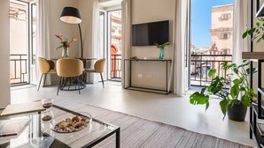 A modern living room with a glass coffee table, a dining area with chairs, a TV mounted on the wall, and a balcony with a view of the city. - Casa San Lio (Noto)