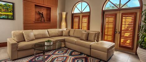 A modern living room with a beige sectional sofa, a glass coffee table, a patterned rug, wooden paneling, a sculpture, and a large window with shutters.